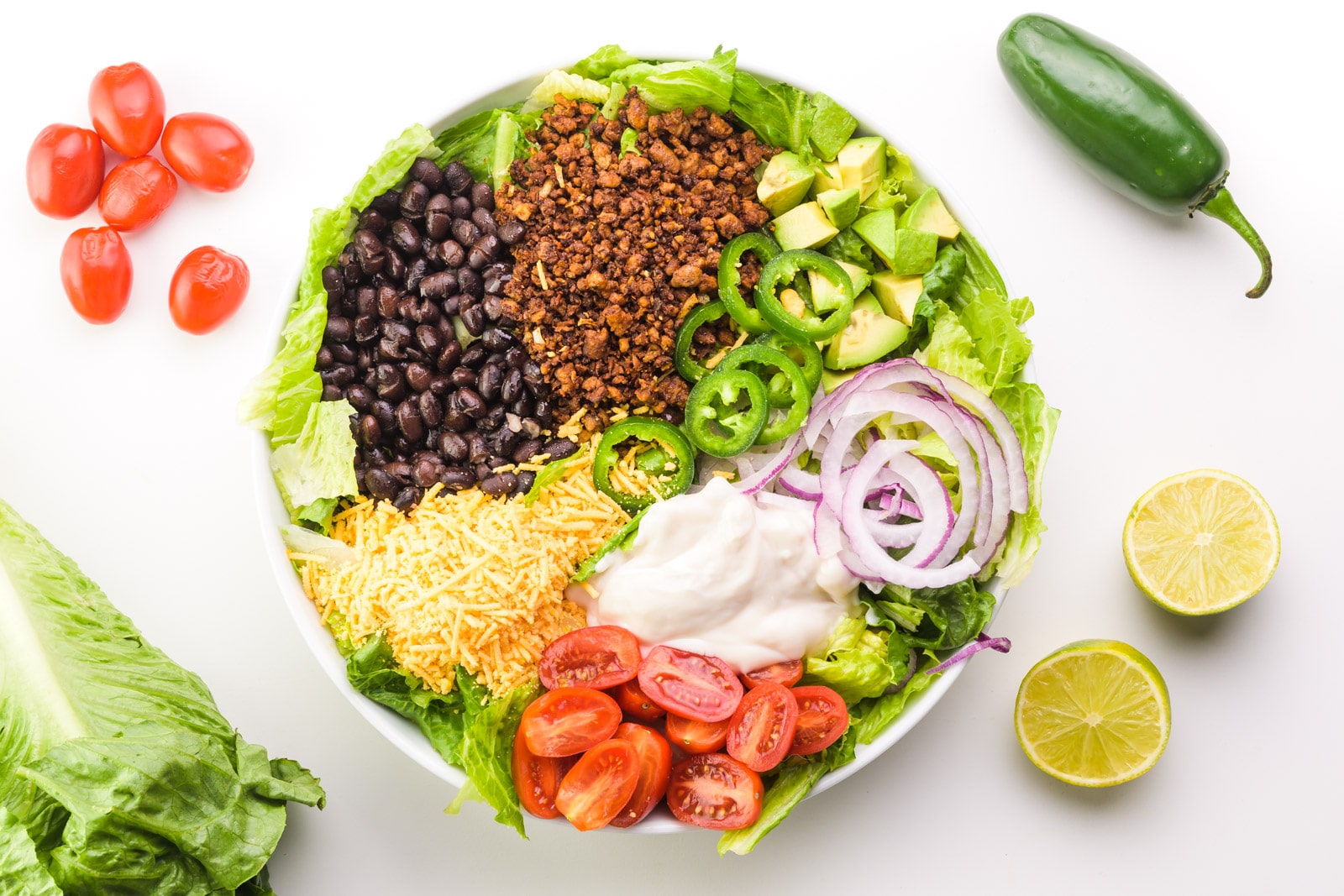 Looking down on a taco salad with lots of veggie toppings, such as red onions, cherry tomatoes, and more. There are sliced limes, a jalapeño, red cherry tomatoes, and more lettuce situated around the bowl.