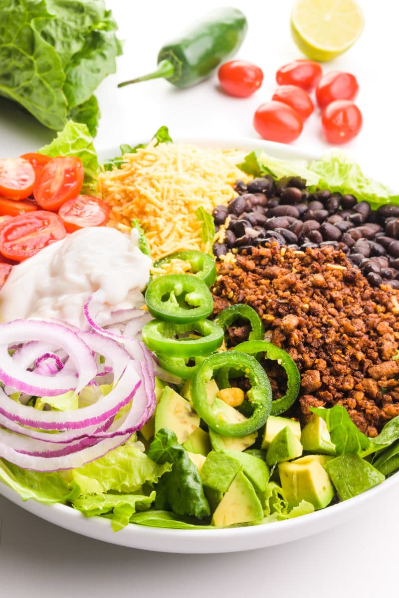 A vegan taco salad is topped with veggie taco meat, black beans, sliced jalapeños and more. There is more ingredients in the background, such as cherry tomatoes and lettuce.