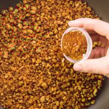 A hand holds a small bowl of spices pouring it over a skillet full of veggie crumbles.