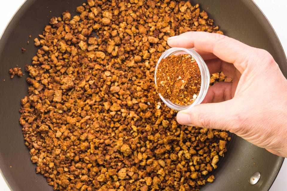A hand holds a small bowl of spices pouring it over a skillet full of veggie crumbles.