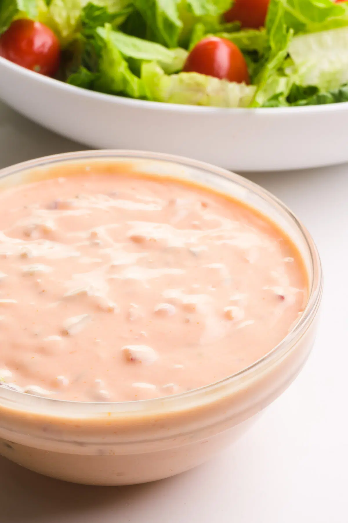A bowl is full of Thousand Island Dressing and sits in front of a salad with greens and cherry tomatoes.