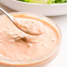 A spon full of vegan thousand island dressing hovers over a bowl full of the dressing. It sits in front of a bowl of salad, barely visible in the background.