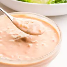A spon full of vegan thousand island dressing hovers over a bowl full of the dressing. It sits in front of a bowl of salad, barely visible in the background.