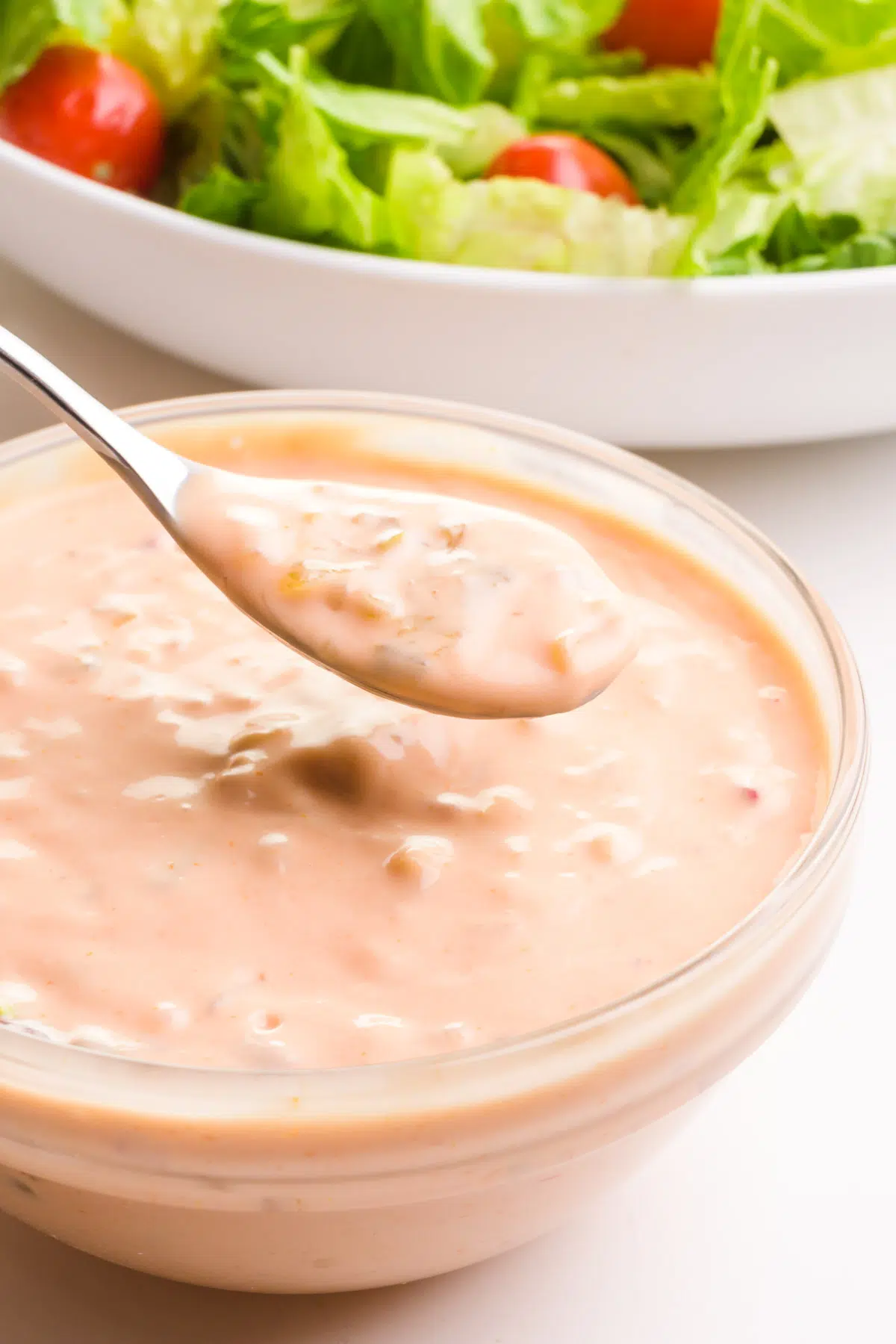 A spoon holds dressing and hovers over a bowl full of the the same creamy dressing. There's a bowl with salad in the background.