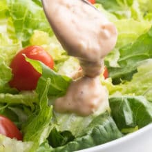 A spoon drizzles vegan thousand island dressing over a bowl of crispy greens and cherry tomatoes.