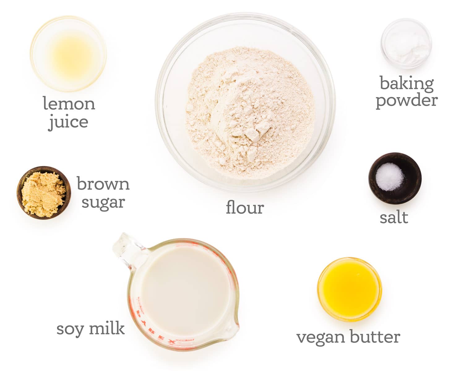 Ingredients are laid out on a white table. The labels next to each ingredient reads, flour, baking powder, salt, vegan butter, vegan milk, brown sugar, and lemon juice.