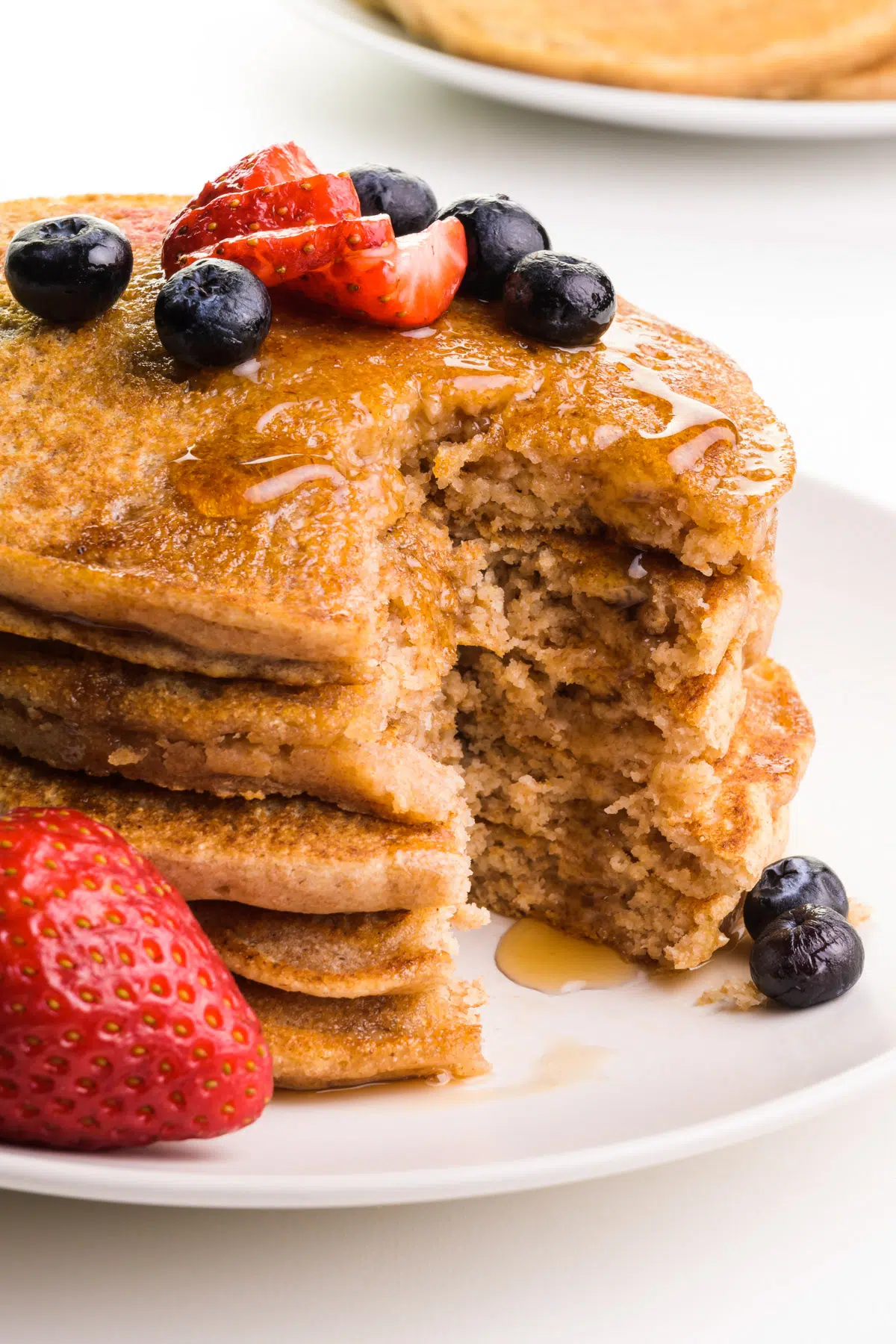 A stack of pancakes has a bite taken out. There's fresh fruit on top and the sides and maple syrup has been drizzled over the top.