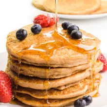 Syrup is being poured on a stack of whole wheat pancakes. The stack has fresh fruit on top and around it. There's a plate with more pancakes in the background.