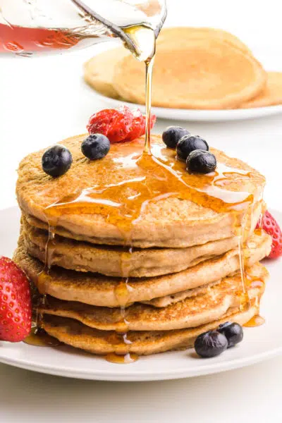 Syrup is being poured on a stack of whole wheat pancakes. The stack has fresh fruit on top and around it. There's a plate with more pancakes in the background.