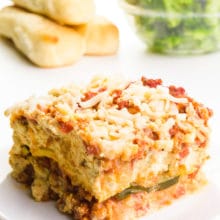 A slice of zucchini lasagna sits on a plate. There are breadsticks in the background and a bowl of steamed broccoli.