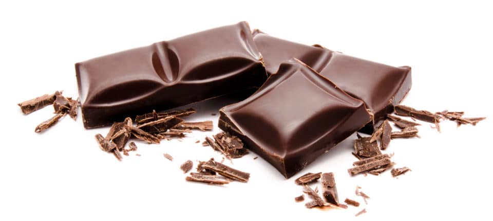 Several pieces of dark chocolate sit on a white counter with flakes of chocolate around them.