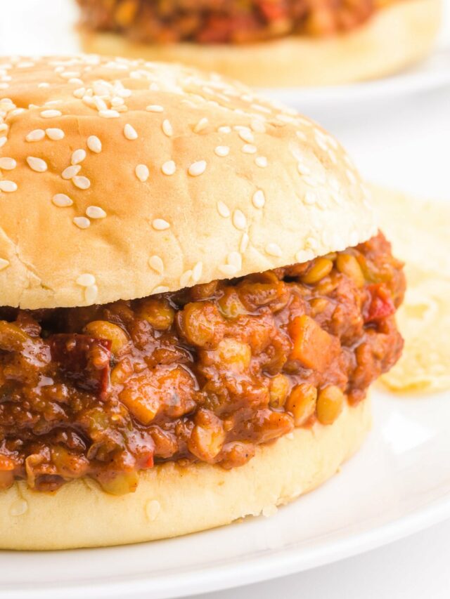 A bun holds lentil sloppy joes on a plate next to potato chips. There's another plate with the same sandwich in the background.