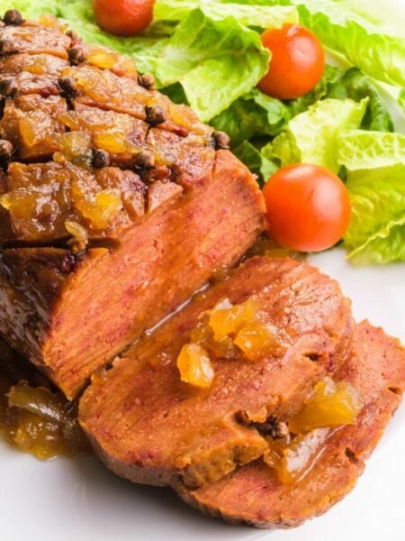 Looking down on a vegan ham with slices cut out. There is pineapple sauce on top of the loaf and on the slices. There is a salad with cherry tomatoes on the back side of the plate.