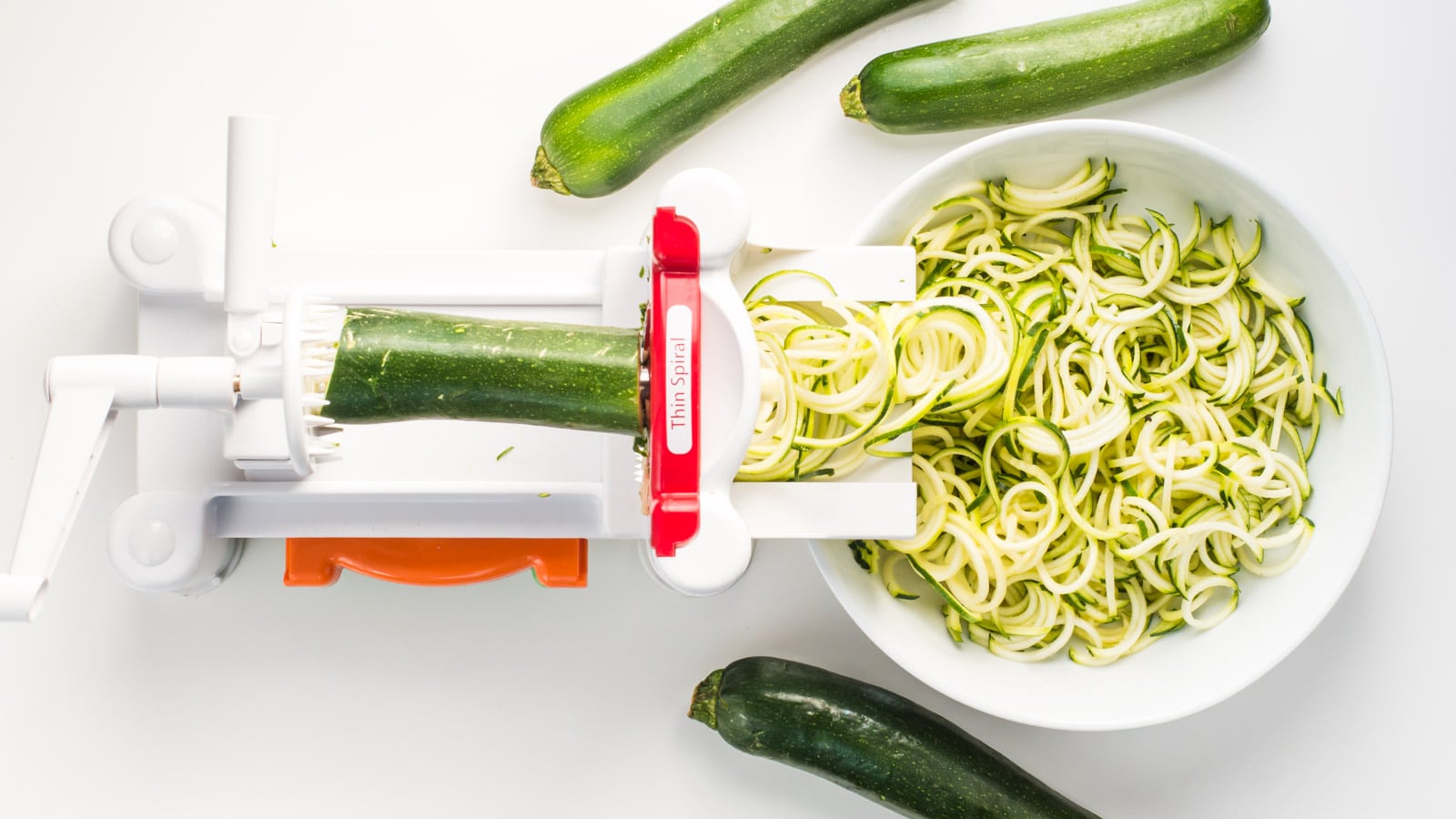 A spiral noodle maker has a zucchini in it and is being used to make zoodles (zucchini noodles). There are 3 whole zucchinis next to the tool.