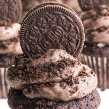 A chocolate cupcake has a generous portion of frosting and an Oreo cookie on top. There are more cupcakes like it in the background. The banner text at the top reads, Vegan Oreo Frosting.