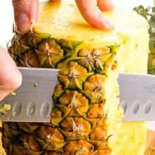 A fresh pineapple has both ends cut off and is sitting tall on a cutting board. A hand holds it steady on top while a knife cuts off part of the skin. The text at the top reads, How to Cut A Pineapple.