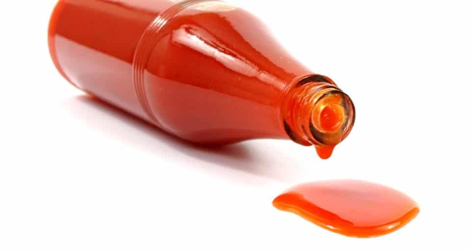 A bottle of hot sauce is on its side, spilling a small pool of sauce on a white counter.