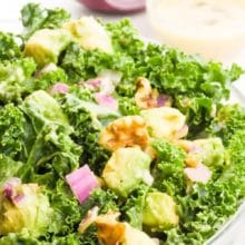 A large bowl holds kale salad, showing curly green kale, chopped walnuts, red onions, and bits of green avocados. There's more of the dressing and a red onion in the background. The text at the top of the image reads, Tahini Dressing Kale Salad.