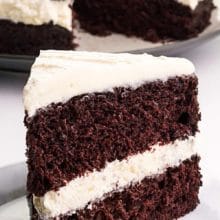 A slice of chocolate cake with white frosting and filling sits on a plate. The rest of the cake is behind it. The text at the top of the image reads, Suzy Q Cake with lots of fluffy filling.