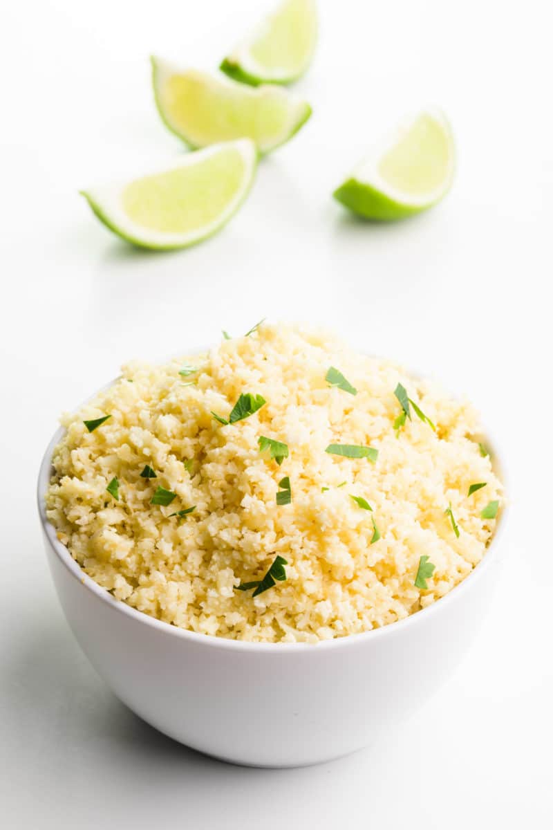 A bowl of cauliflower rice sits on a white table in front of lemon slices.