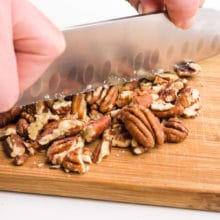 A hand holds a knife, chopping pecans on a cutting board.