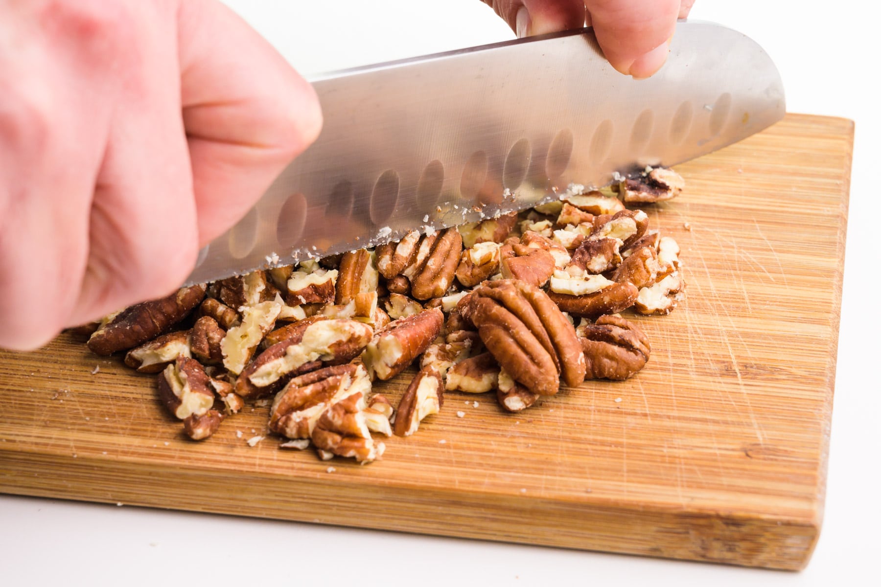 A hand holds a knife, chopping pecans on a cutting board.