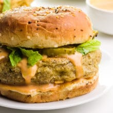 A fried tofu sandwich sits on a plate. It has a creamy sauce, pickles, and lettuce on top.