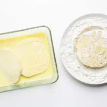 A rectangle baking dish has a soy milk mixture in it and two tofu patties. A plate sits next to it with a flour mixture and another tofu patty in that.