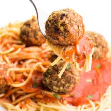 A fork holds a lentil meatball on top of spaghetti and red sauce. It hovers over a plate of more of the dish.