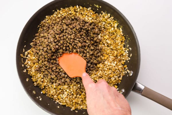 A hand holds a spatula, reaching into a skillet with lentils and cooked onions.