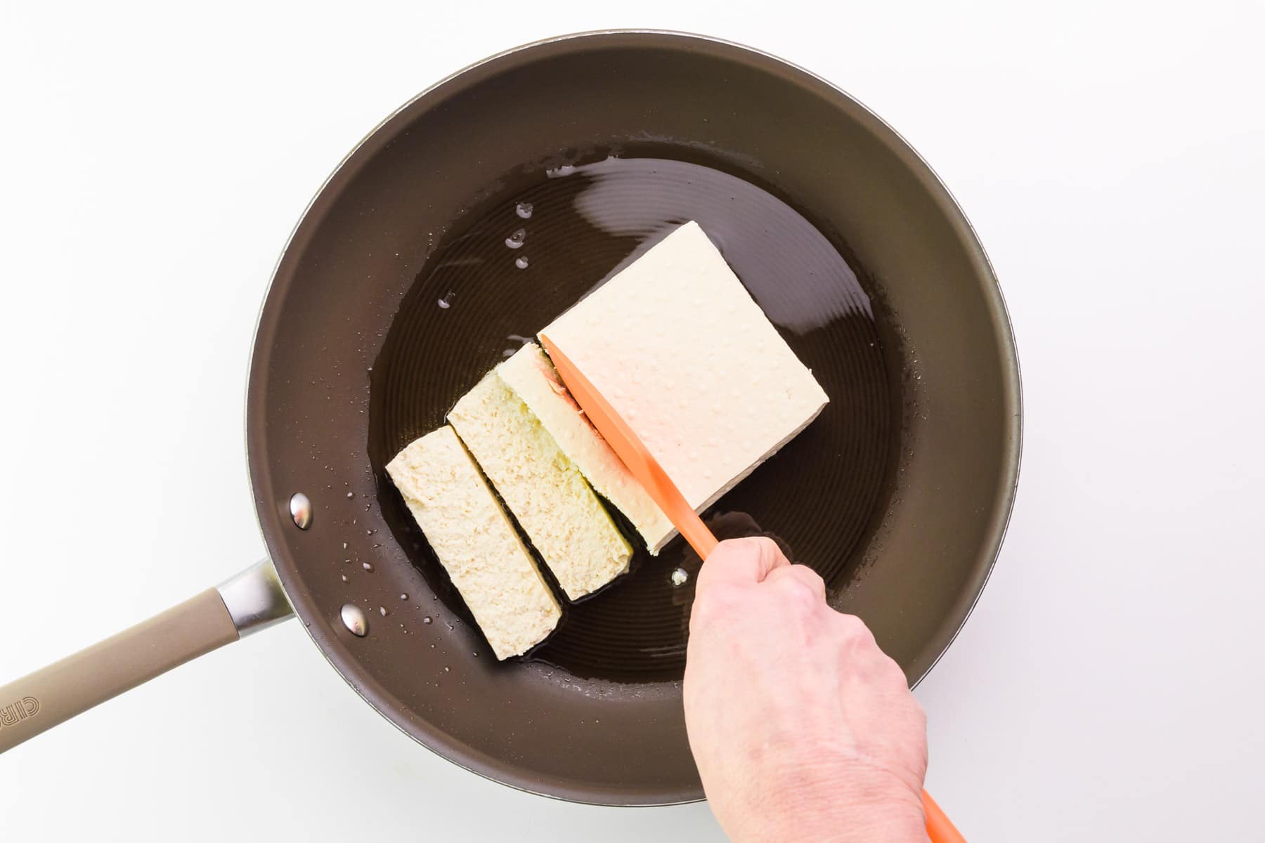 A hand holds a spatula, breaking tofu in a skillet into smaller pieces.