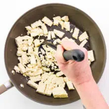 A hand uses a potato masher to mash tofu in a skillet.