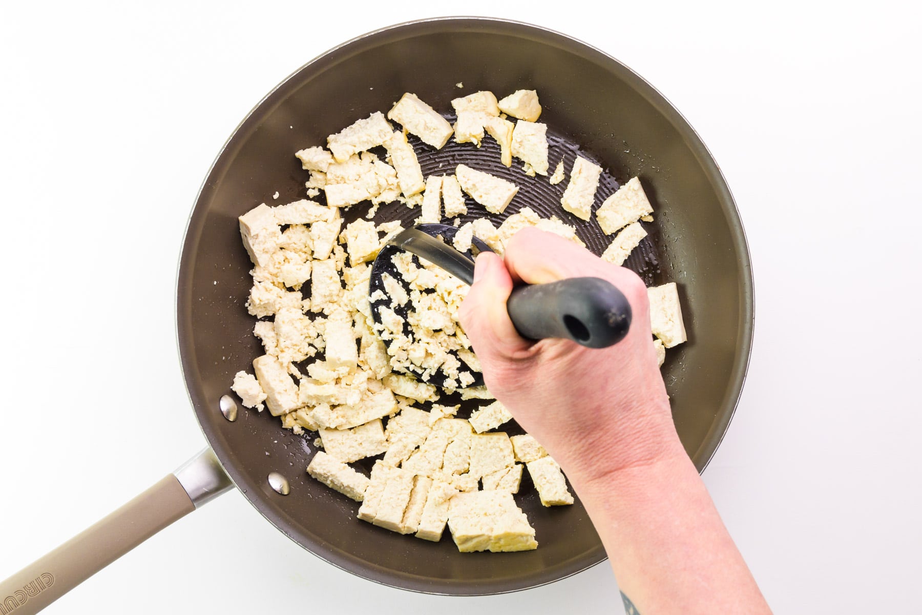 A hand uses a potato masher to mash tofu in a skillet.