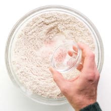 A hand holds a bowl sprinkling pink salt over a bowl with flour.
