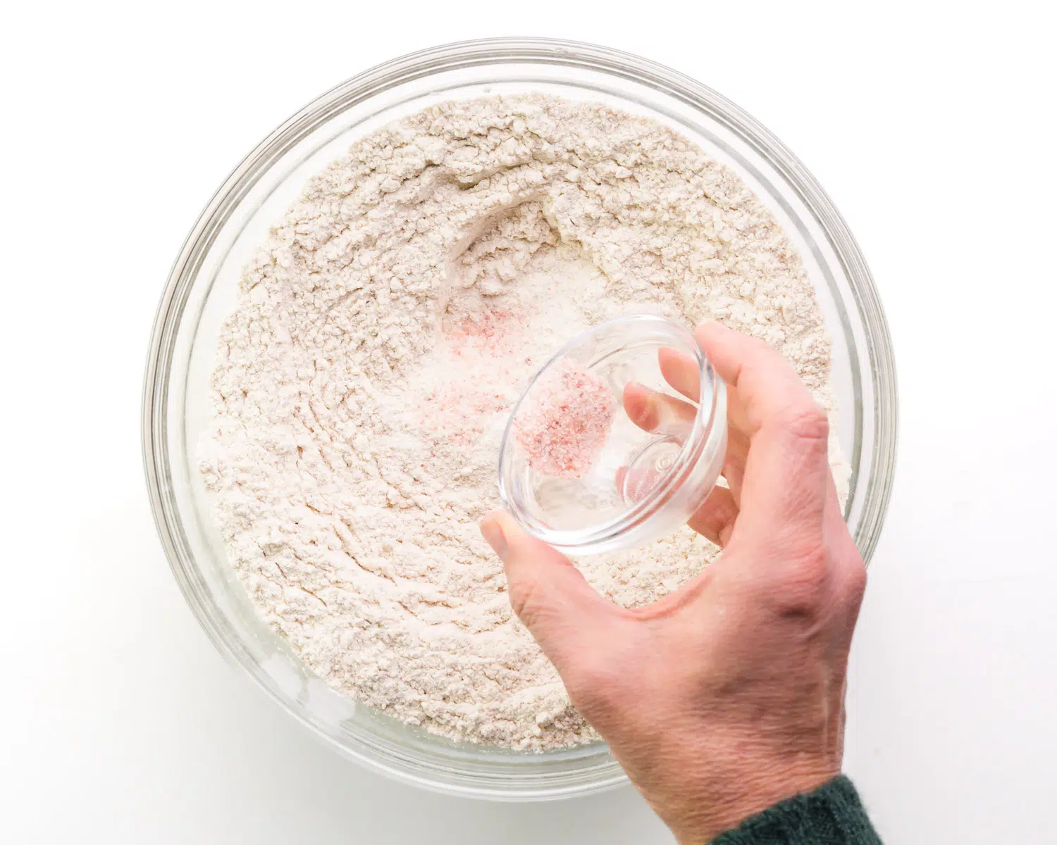 A hand holds a bowl sprinkling pink salt over a bowl with flour.