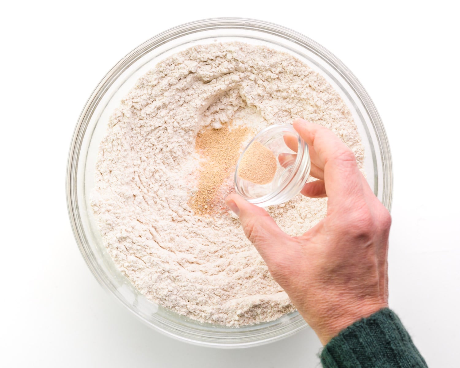 A hand holds a bowl with dry yeast, sprinkling it over a bowl of flour.