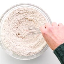A hand holds a whisk, stirring a flour mix in a bowl.
