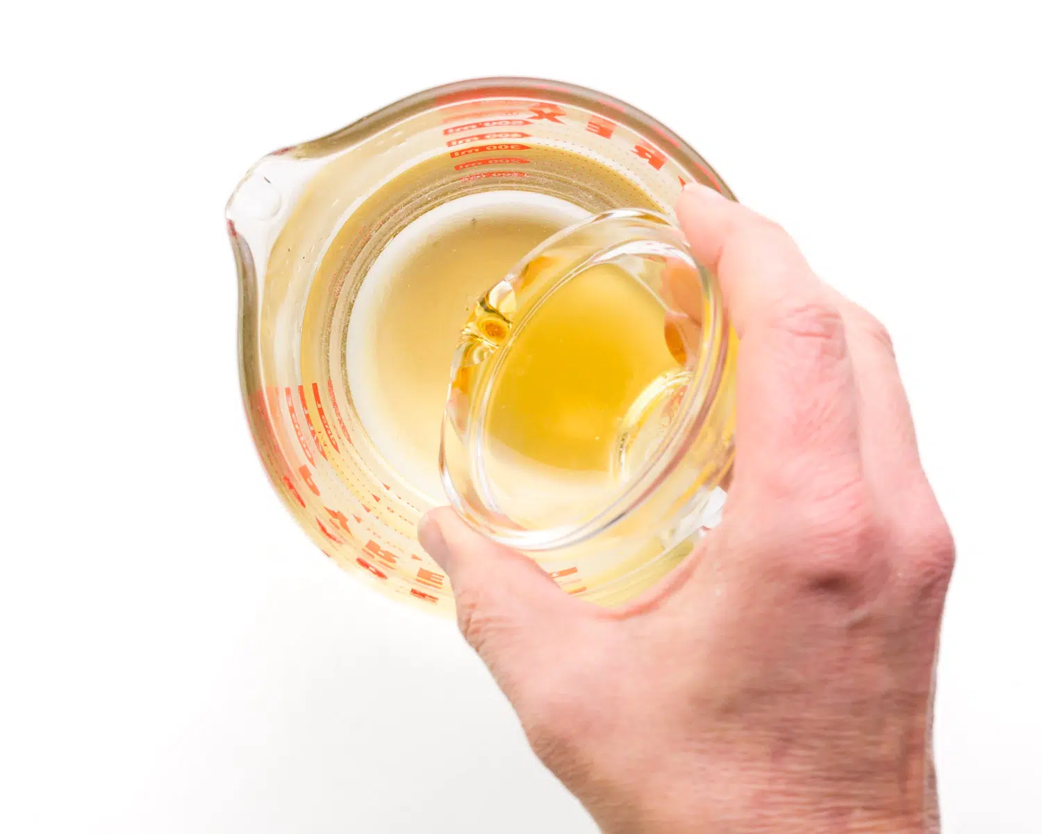 A hand holds a small bowl of maple syrup, pouring it into a glass measuring cup with water.