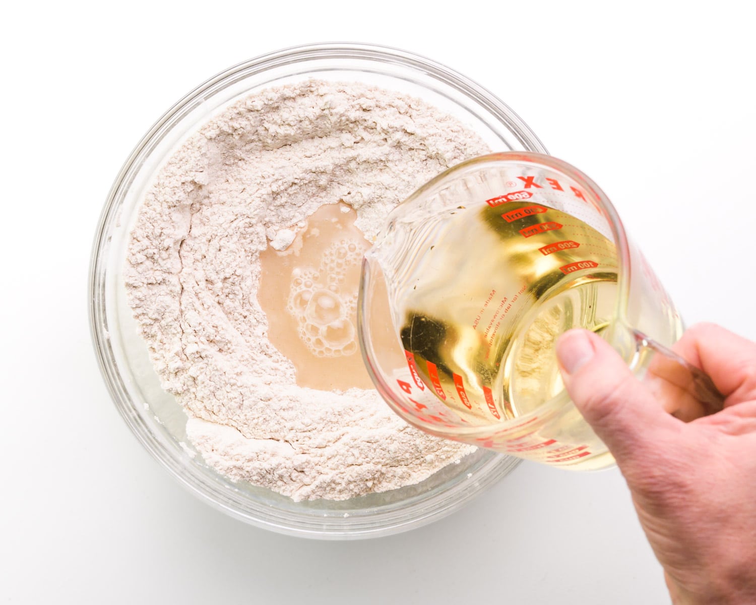 A hand holds a glass measuring cup, pouring a liquid ingredient into a bowl of flour.