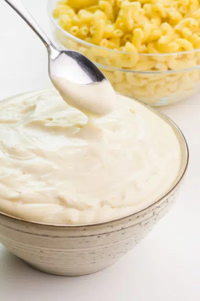 A spoon hovers over a bowl of vegan béchamel sauce. The spoon has some sauce in it and it is drizzling into the bowl. There's a bowl of macaroni in the background.