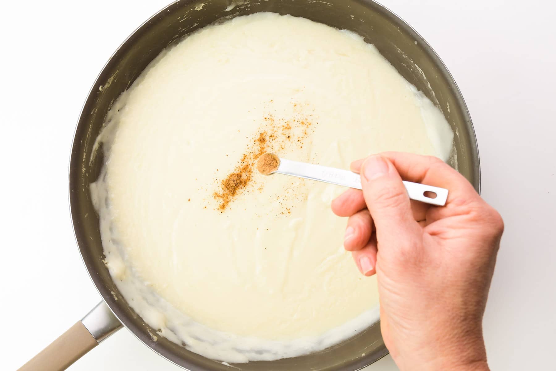 A hand holds a measuring spoon, sprinkling ground nutmeg into vegan white sauce in a skillet.