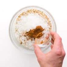 A hand holds a bowl of ground cinnamon, pouring it into a bowl with other ingredients like oats and coconut flakes.