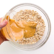 A hand holds a bowl of peanut butter, pouring it into a bowl with an oats mixture.