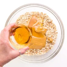 Maple syrup is being poured into a bowl with peanut butter and oats.