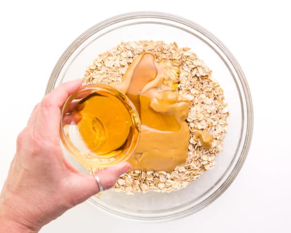 Maple syrup is being poured into a bowl with peanut butter and oats.