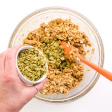 A hand holds a bowl of pumpkin seeds, pouring them into a bowl with cookie dough.