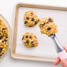 A hand holds a cookie dough scoop full of cookie dough over a baking pan with more cookie dough balls on it. There's a bowl with more dough on the side.