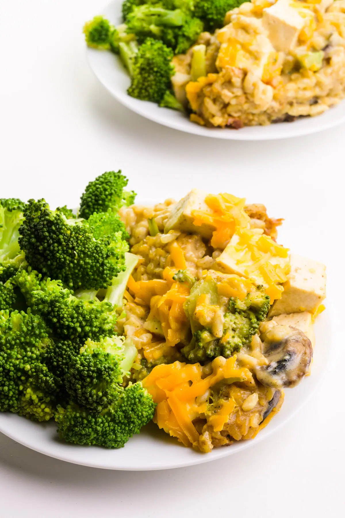 A portion of casserole sits on a plate next to steamed broccoli. There's another similar plate in the background.