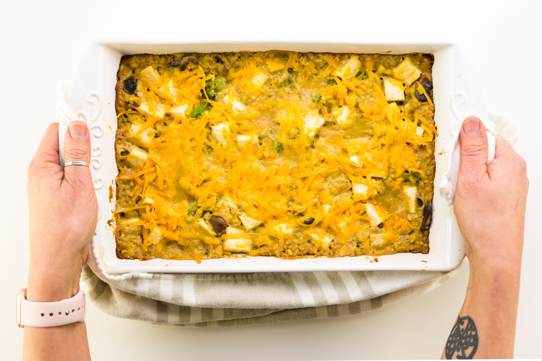 Hands hold both ends of a baking dish with a kitchen towel. The dish holds a cheesy casserole.