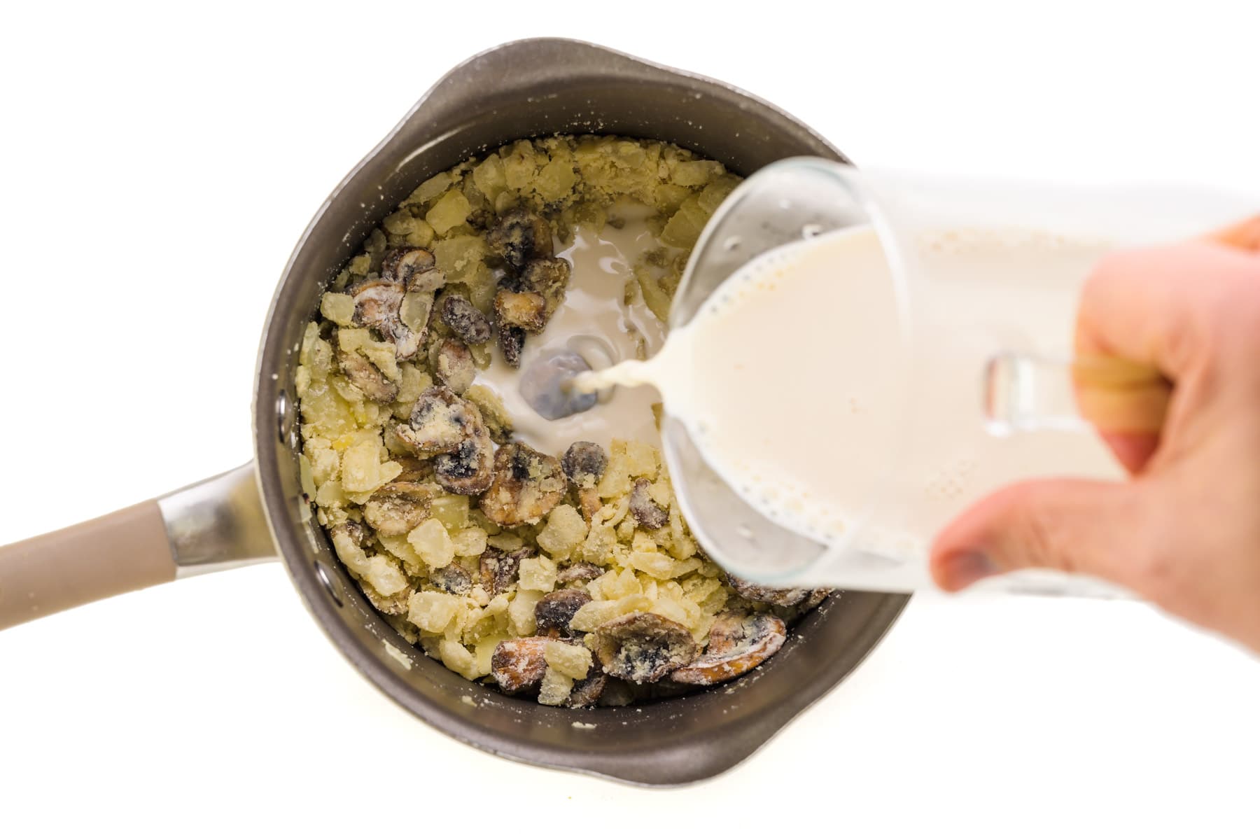 Plant-based milk is being poured into a saucepan with mushrooms and a roux.